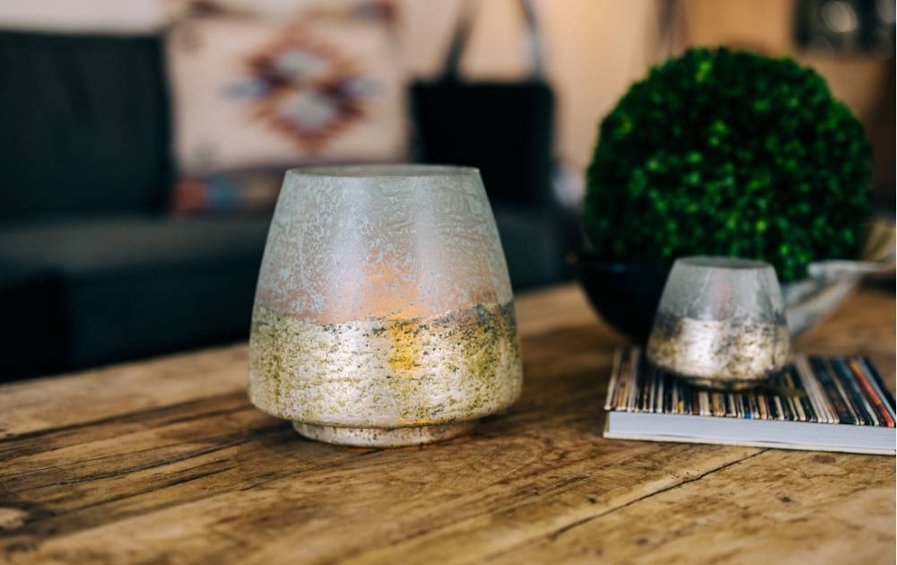A frosted candle holder with a golden base sits on a wooden table beside a stack of magazines, a small artificial green plant, and a gray couch with a patterned cushion in the background.