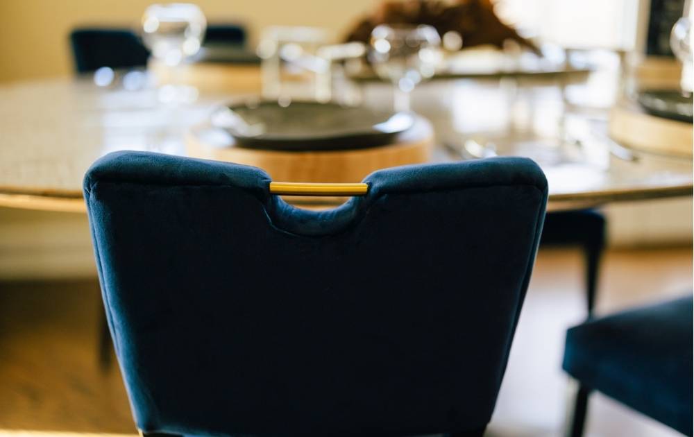 Close-up of a dark blue velvet dining chair with a gold handle at a round table, set with glassware and plates, in a well-lit dining room.