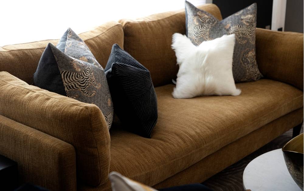 A modern living room with a blue velvet sofa decorated with abstract-patterned and green pillows, a round side table with a marbled vase and plant, and abstract art on taupe, slatted walls.