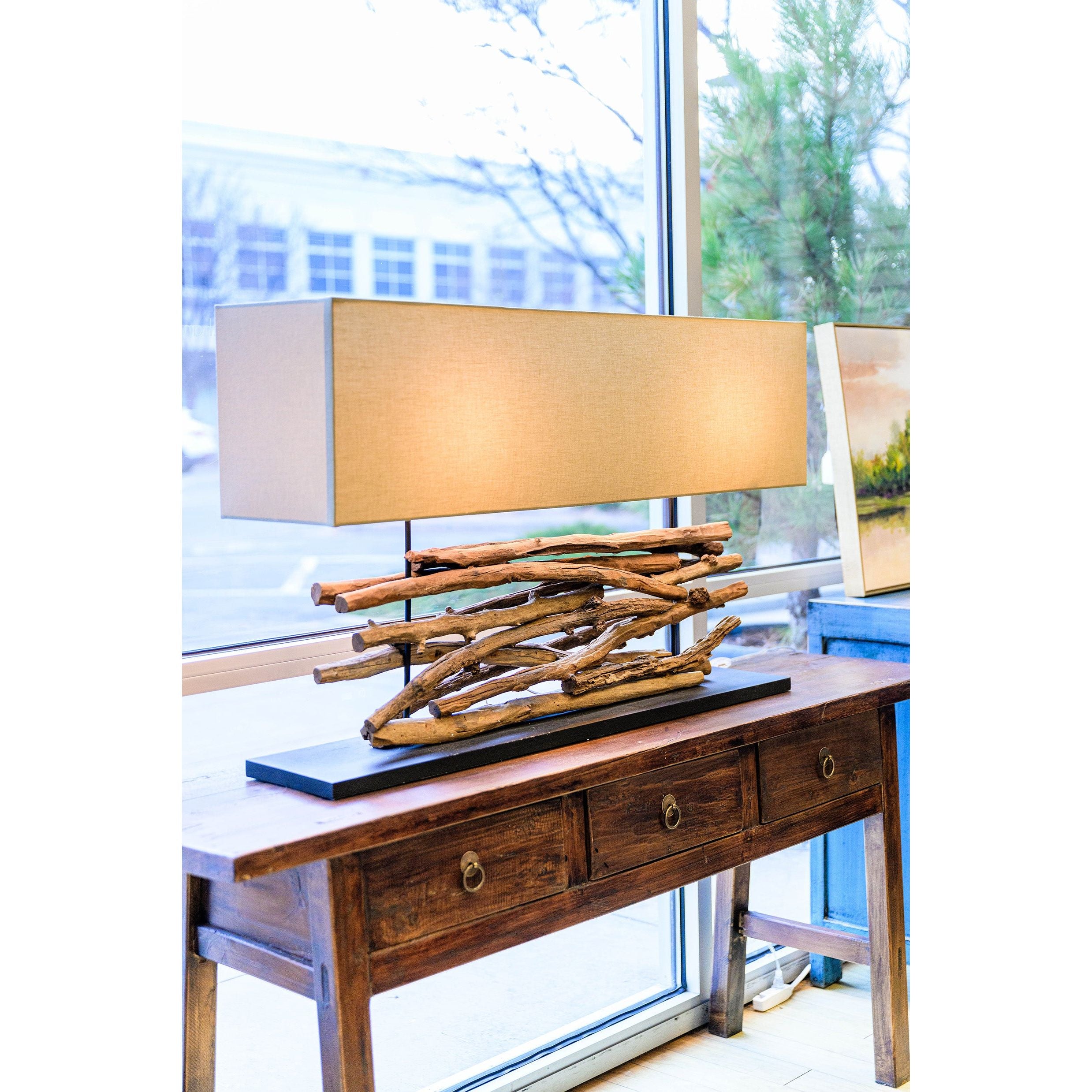 A rectangular lamp with a beige shade and a base of entwined natural branches rests on the Tigris Console Butcher Table, made from reclaimed wood and featuring two storage drawers with round handles; a window and painting are in the background.