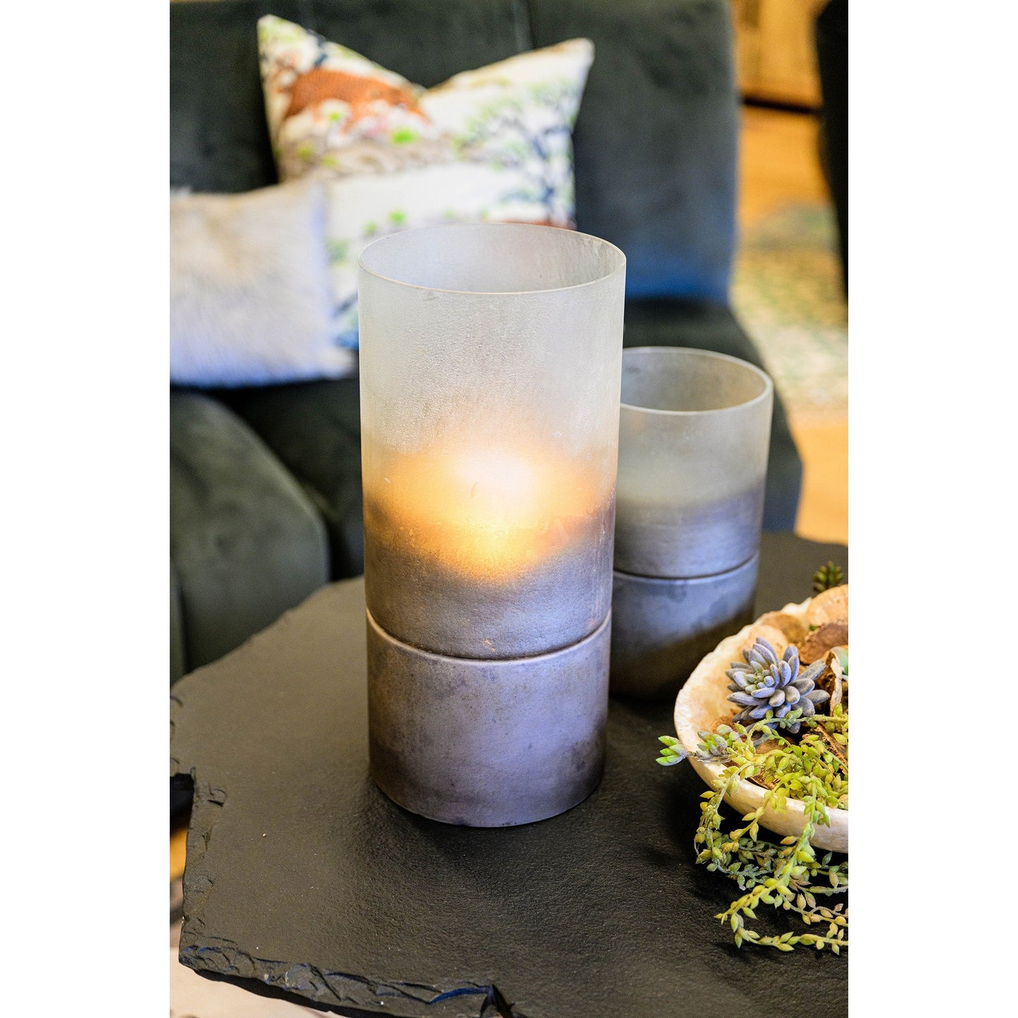 The Clara Metallic Smoke Hurricane, with a lit candle inside, elegantly sits on a dark textured table. In the background, a soft green couch with floral pillows complements the scene, while a small bowl of succulents enhances the transitional home decor ambiance.