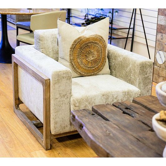 A cozy Emma Accent Chair with a beige velvety fabric and wooden frame in a cafe setting, adorned with a round, decorative cushion with a sunburst pattern. A rustic wooden coffee table sits in front, embodying rustic elegance.