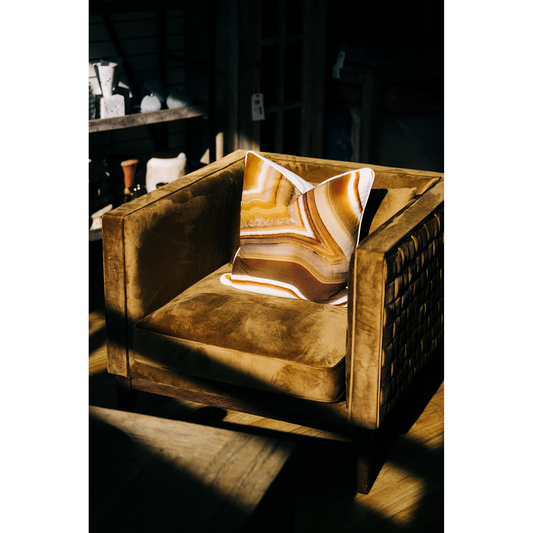 An elegant Amber Accent Chair with a patterned cushion, bathed in dappled sunlight, set against a dimly lit, rustic background, reflecting a serene and cozy ambiance.