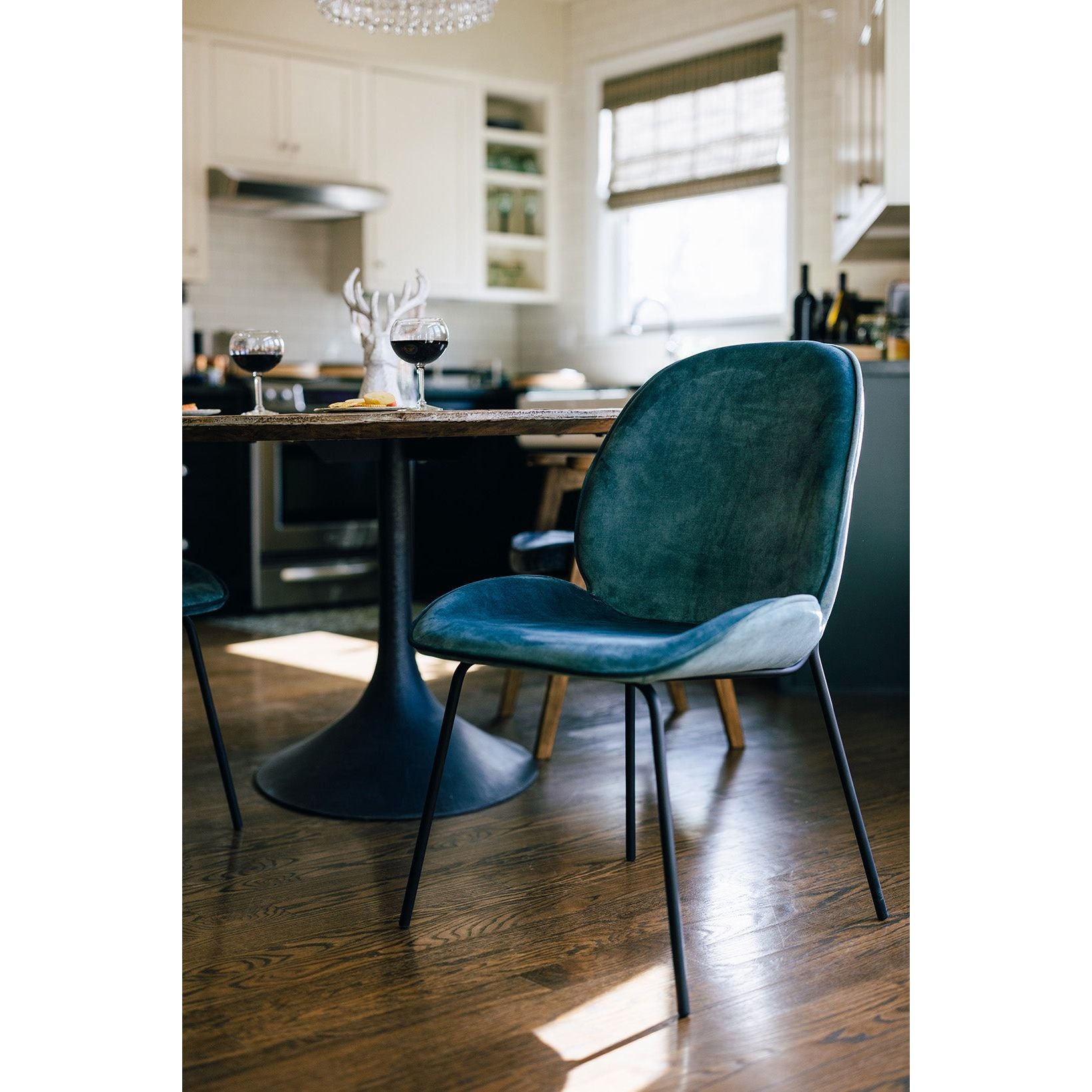 A modern kitchen features a black round pedestal table with two glasses of red wine and a decorative white antler sculpture on top. A King Dining Chair, Smoked Turquoise sits in the foreground on a wooden floor, while bright natural light fills the space.