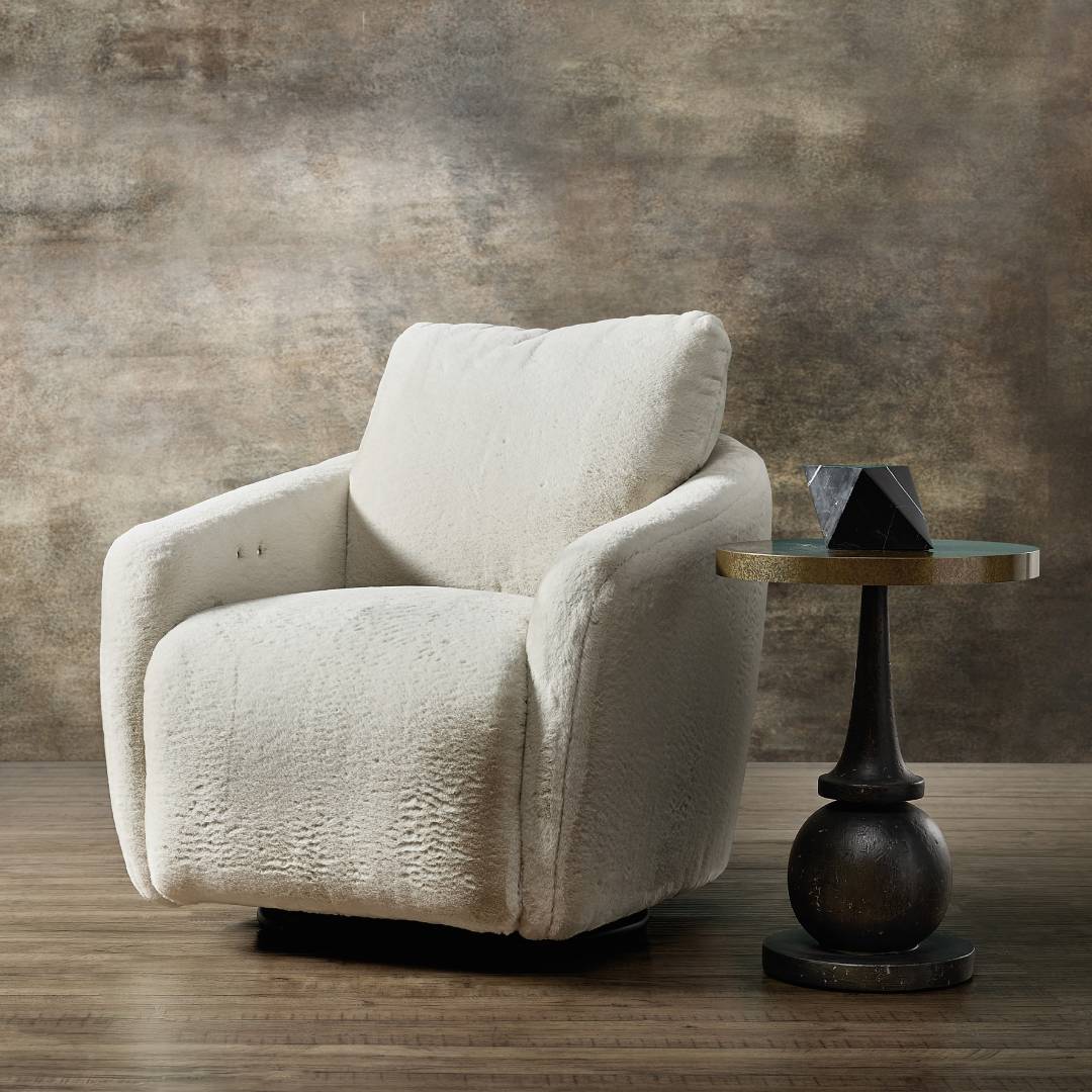 The Arista Swivel Chair with Power Footrest, in plush white faux fur, sits on a wooden floor next to a round, gold-trimmed side table and black geometric decor, set against a textured earth-toned wall.