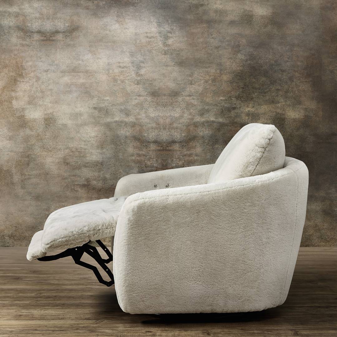 A side view of the Arista Swivel Chair with Power Footrest, upholstered in plush white textured fabric, showcased on a wooden floor against a rustic mottled brown wall.