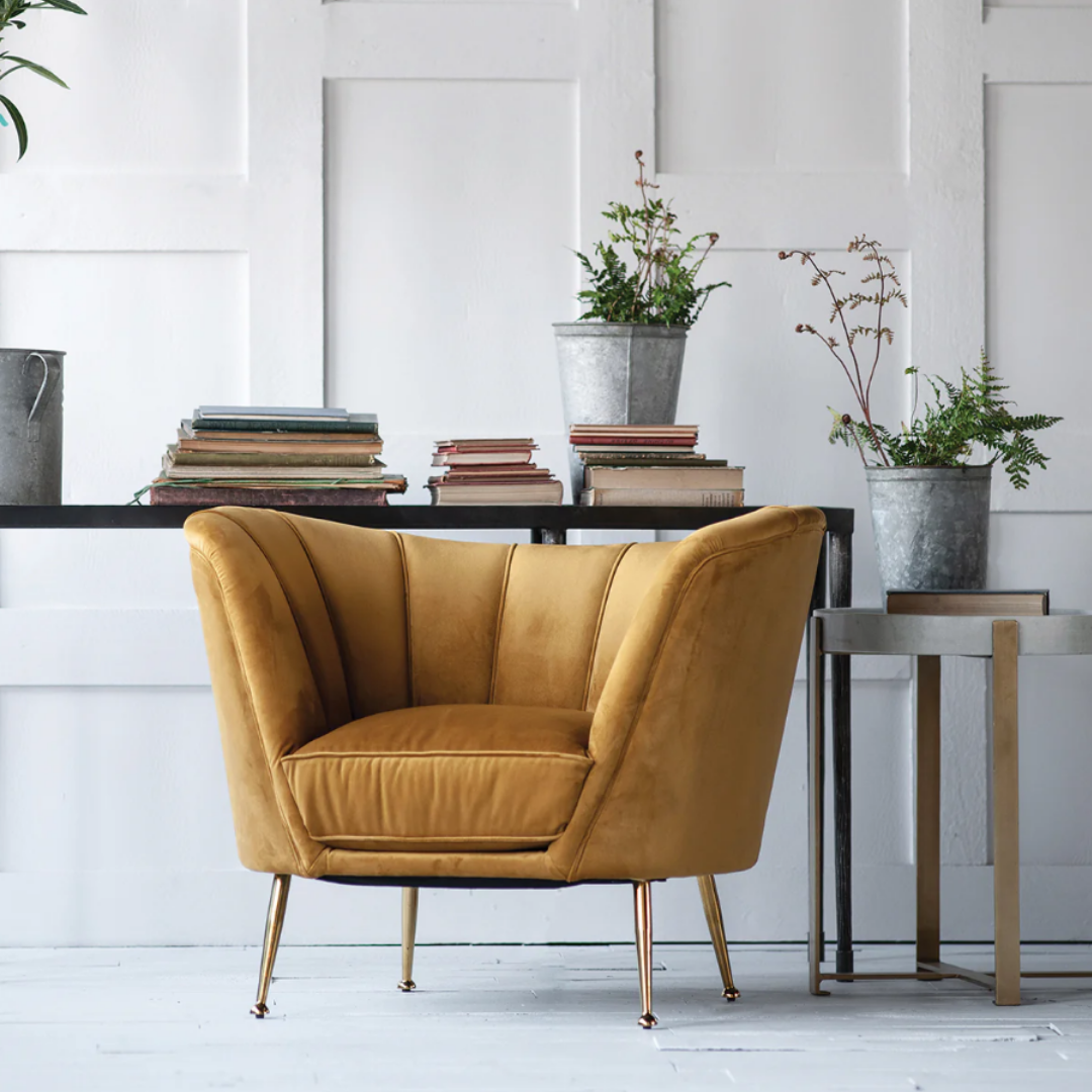 The Calvierre Channel-Tufted Velvet Chair in cozy mustard yellow with metal legs rests on a light wooden floor. A dark table behind it holds books and metal planters, while a small square side table with a plant enhances the modern decor against the white paneled wall.