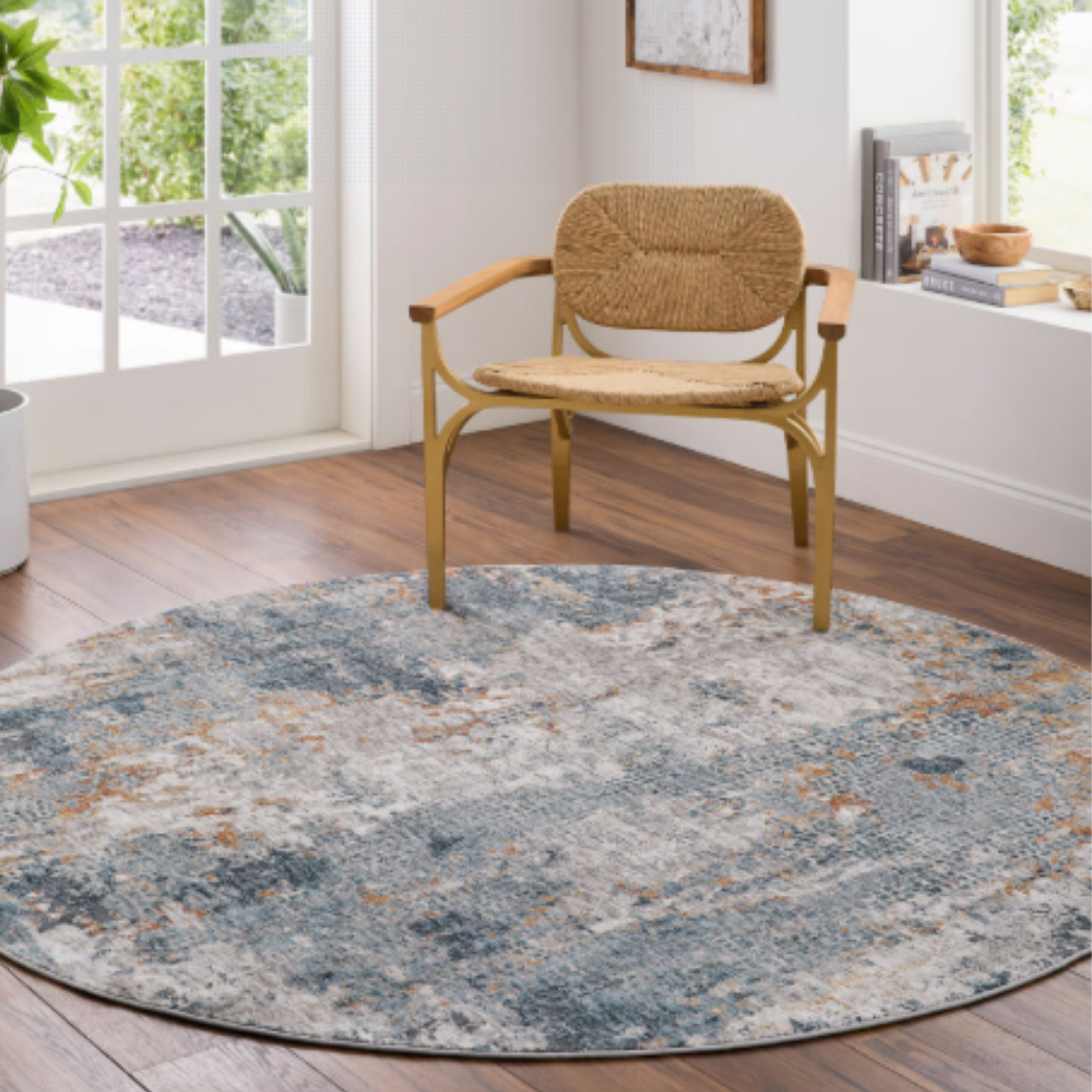 The Candice Machine Woven Rug, featuring blue, gray, and beige abstract patterns, sits on a wooden floor beneath a wicker chair. Large windows and white walls complete this bright, modern room with minimal decor.