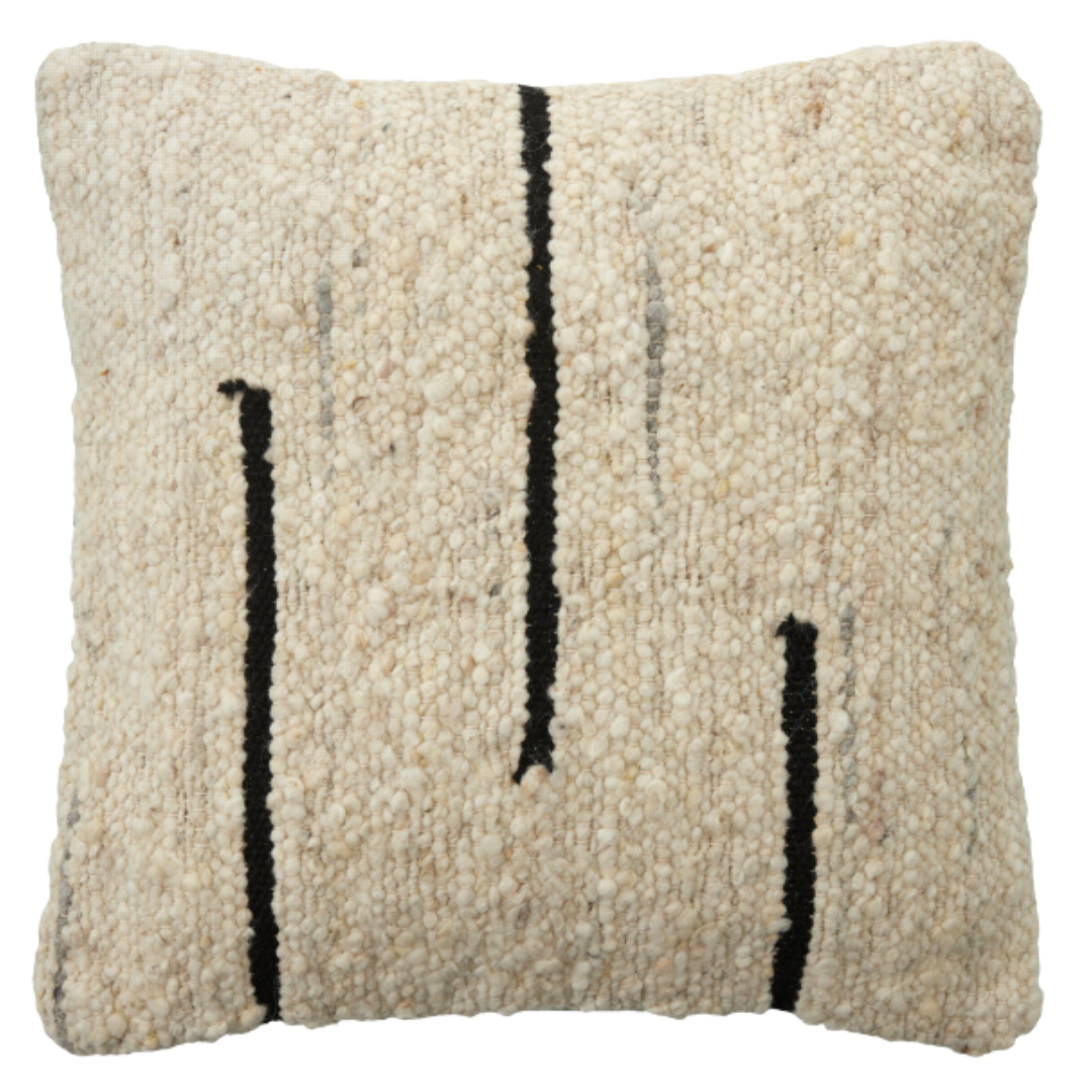 The Dillon Wool Pillow is a square, cream-colored cushion with a soft, textured weave and three uneven vertical black lines on the front, offering a modern, minimalist look. Plush and comfortable, it resembles a cream and black woven rug.