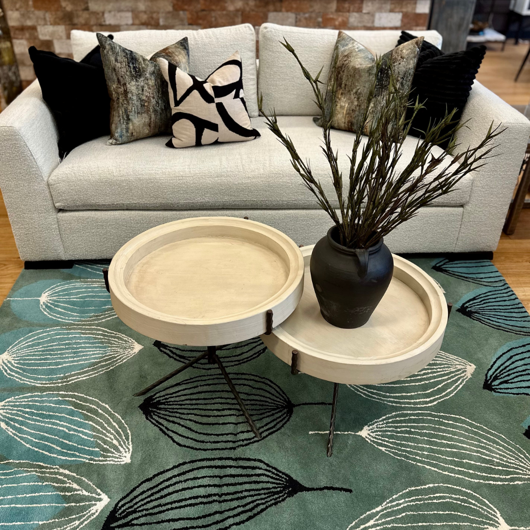 A modern living room with the Dresden Performance Fabric Customizable Sofa, black and patterned pillows, a teal leaf-patterned rug, round wooden nesting coffee tables, a black vase with tall branches, and a brick accent wall in the background.