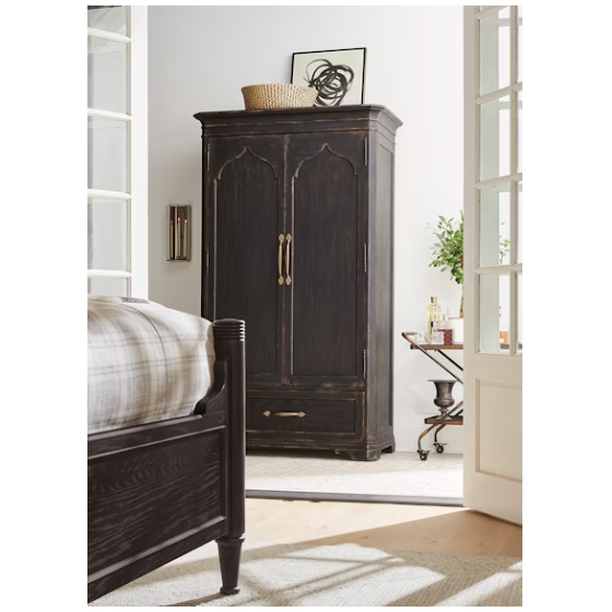 The Florence Wardrobe Cabinet with brass handles stands in a bright bedroom next to a matching bed; atop the wardrobe are a woven basket and abstract art. A bar cart and plant are near the French doors in the softly lit space.