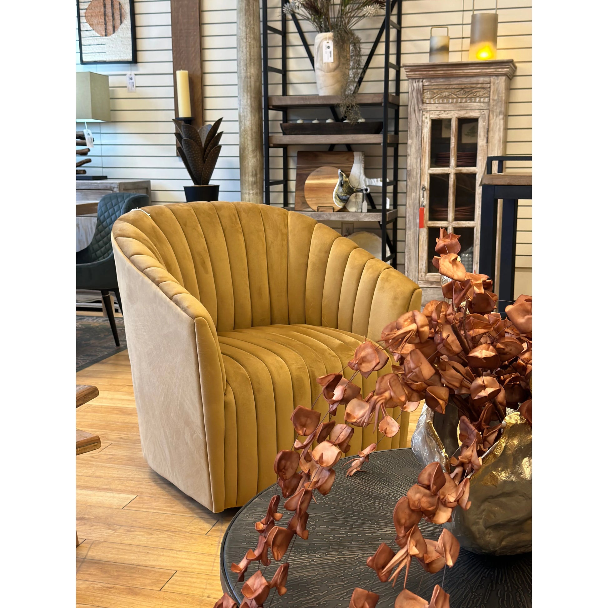 A luxurious Adaline Swivel Accent Chair in mustard yellow polyester with vertical pleats sits in a stylish home decor showroom featuring shelves with various decorative items, and a foreground of shiny copper leaves spilling from a metallic vase.