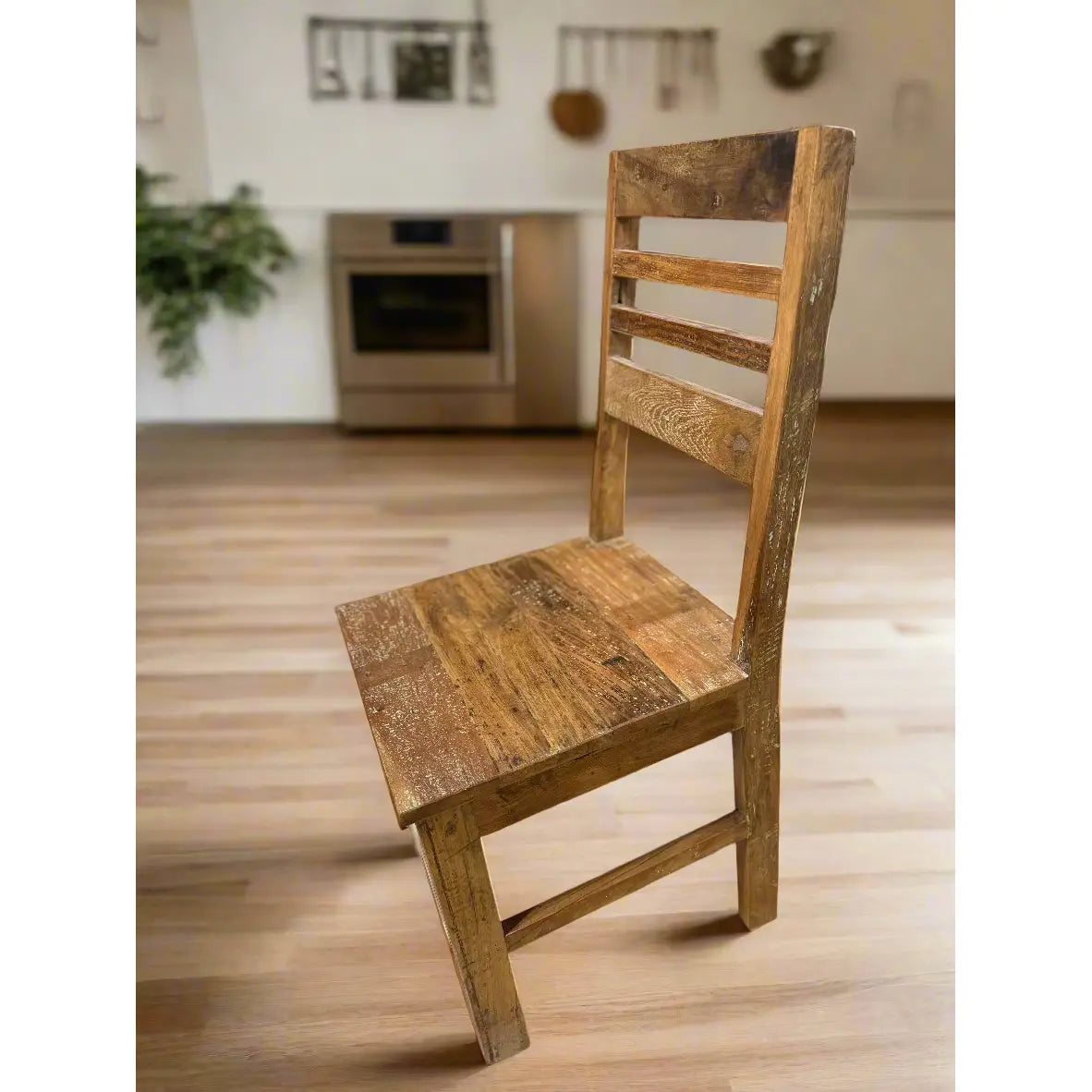 The Wood Dining Chair, handcrafted with rustic charm and a simple design, features a slatted back and sturdy seat. It sits on a light wood floor with a modern kitchen, beige oven, hanging utensils in the background, and a potted plant to the left.