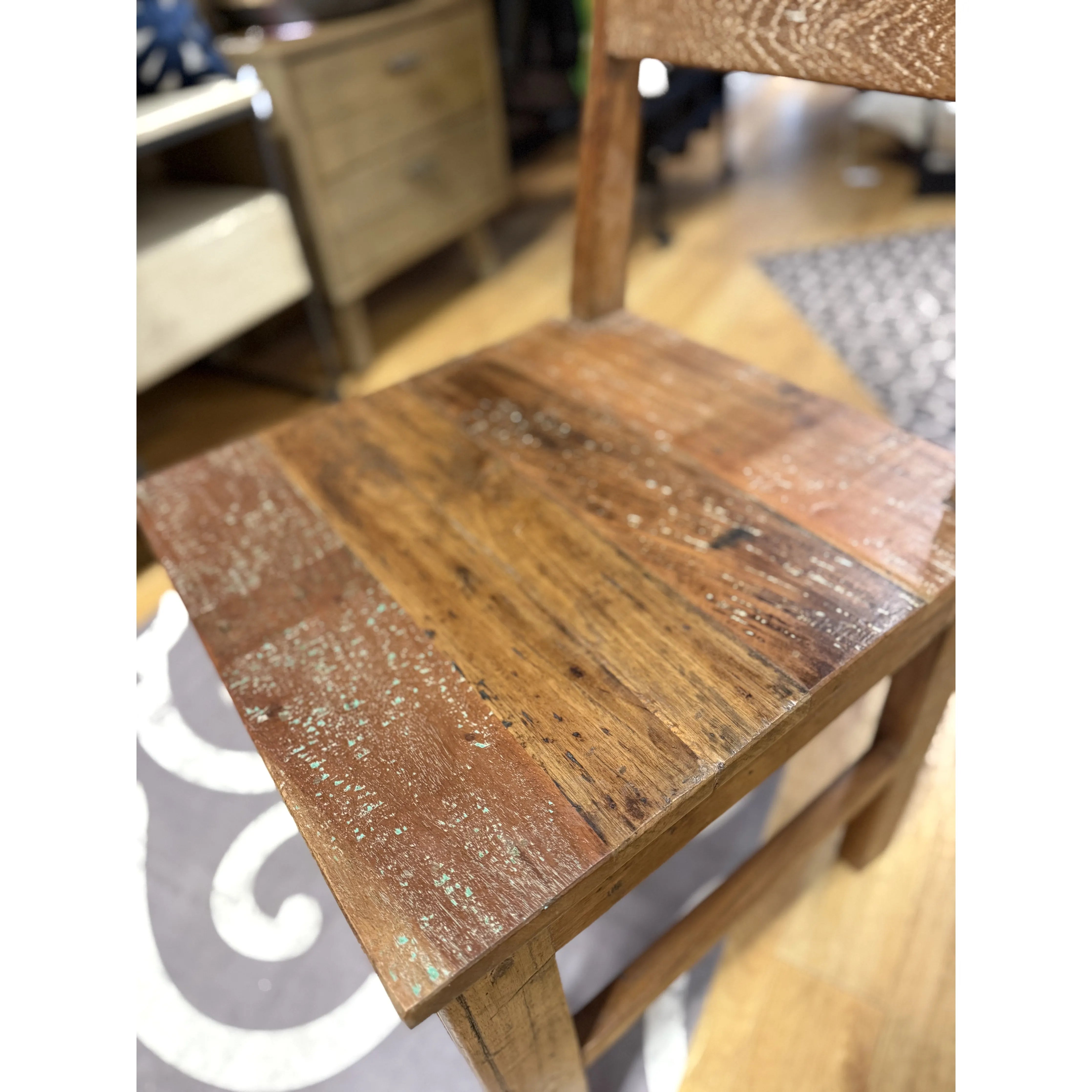 The Wood Dining Chair, featuring a distressed finish and visible wood grain for rustic charm, is the focal point, while blurred in the background are a chest of drawers and a gray and white patterned rug on a wooden floor.