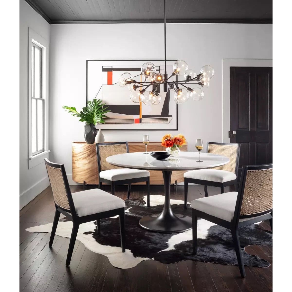Modern dining room with a round white table and four Antonia Cane Armless Dining Chairs with high-performance fabric seating on a black and white cowhide rug. A contemporary chandelier with exposed bulbs hangs above the table. The room features a black modern art piece on the wall and a wooden side cabinet with decor.