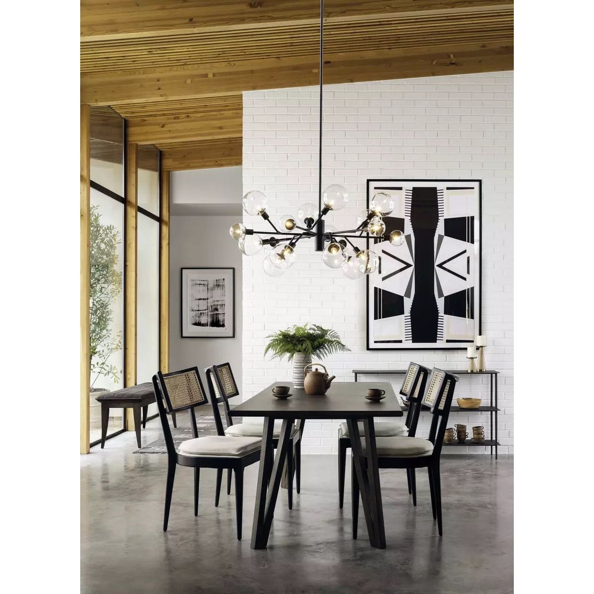 A modern dining room features an ebony-finished nettlewood dining table and four matching Britt Dining Chairs with high-performance linen-blend seating. A large, contemporary chandelier with multiple bulbs hangs above the table. A white brick wall displays black and white abstract art, while a wooden ceiling and large windows bring in natural light.