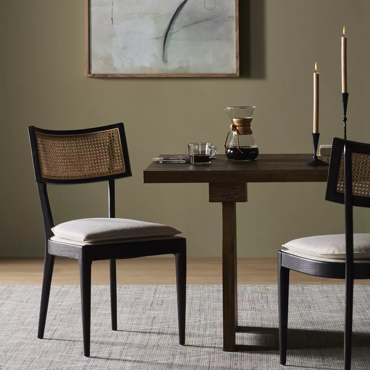 A modern dining area featuring a wooden table with two Britt Dining Chairs. The chairs have woven cane backs and cushioned seats. On the table are a Chemex coffee maker, a small cup, and two lit taper candles. A minimalist abstract painting hangs on the wall behind the table.