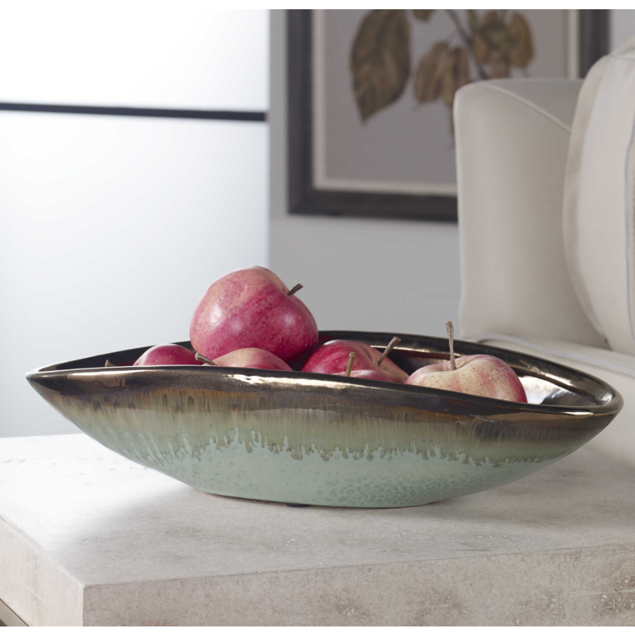 A shallow, earthenware Iroquois Bowl filled with red apples sits on a light-colored marble table. The bowl features a smooth, gradient finish transitioning from mint green glaze at the base to a bronze color at the rim. In the background, there is a portion of a white chair and a framed artwork of leaves.