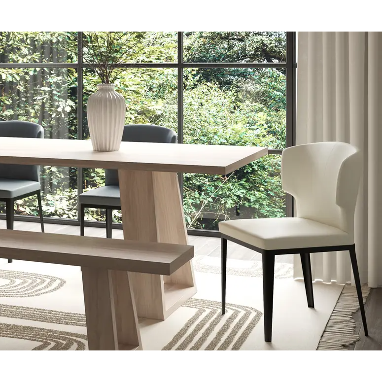 A modern dining room includes a wooden table with a matching bench beside the Randolph Dining Chair, upholstered in white vegan leather. A large textured vase with dried branches adorns the table. Floor-to-ceiling windows reveal lush greenery, and light curtains along with a patterned rug add warmth and texture.
