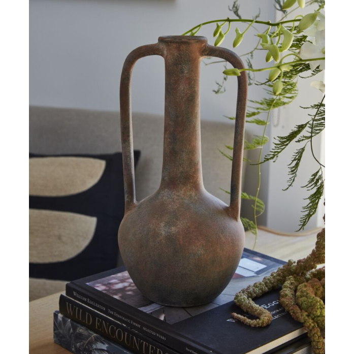 A Tupita Vase with two handles and a narrow neck rests on stacked books atop a wooden table. Behind it, a beige sofa with a black-and-beige cushion and green trailing plants create a warm, inviting atmosphere.