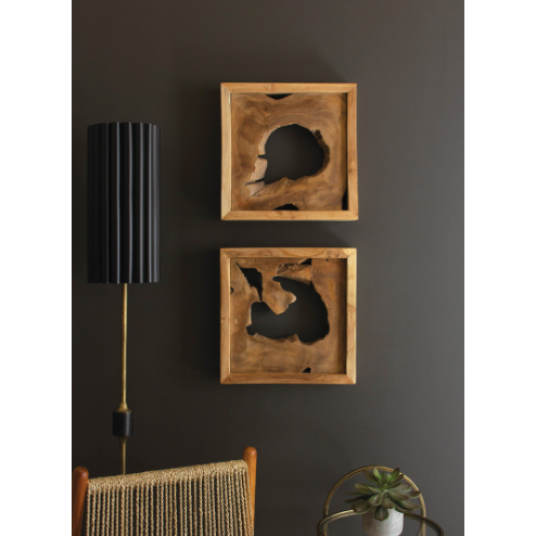A modern interior features a dark wall adorned with two Teak Wall Art pieces, showcasing natural, irregular cutouts. Below the frames is a tall black cylindrical lamp. A woven seat and a small round table with a potted succulent plant are partially visible at the bottom of the image.