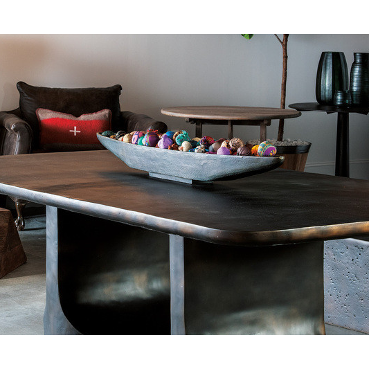 A modern living room scene featuring a large, rectangular coffee table with a minimalist design. Atop the table is a rectangular stone bowl filled with colorful handsewn Textile Balls. In the background, there is a dark armchair adorned with a red and white accented cushion, and shelves displaying decorative vases.