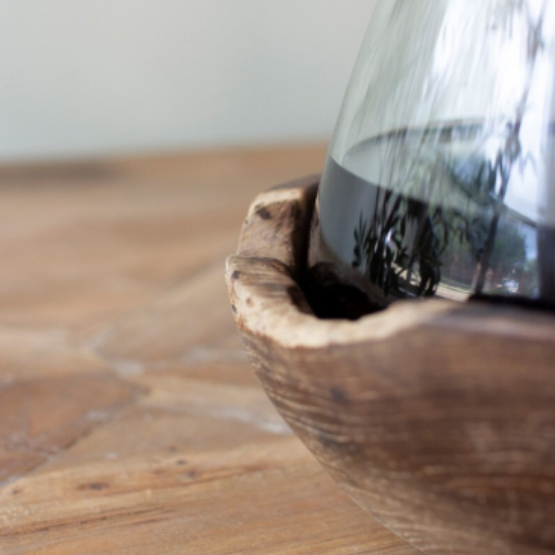 A close-up of the Teakwood & Blown Glass Bottle with dark liquid, nestled in a rustic carved teakwood base on a light wooden surface, with a softly blurred background.