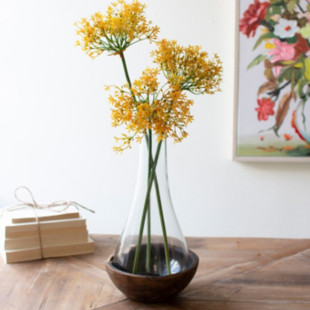 A Teakwood & Blown Glass Bottle holds three tall yellow allium stems on a wooden table. Next to it, a stack of books tied with string sits in front of part of a vibrant floral painting.
