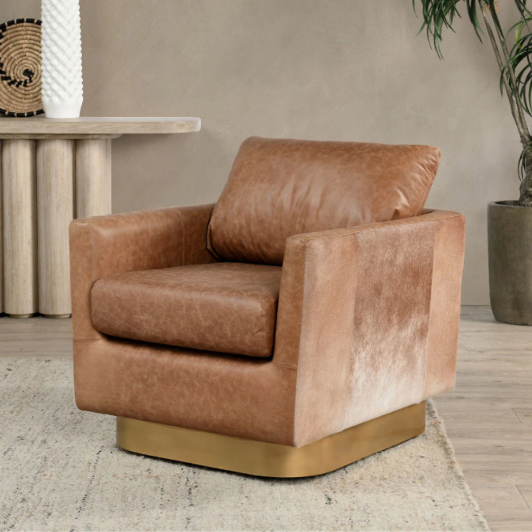 A modern glam scene features a Velancia Hide & Leather Swivel Chair with plush brown top grain leather cushions on a wooden base centered on a beige textured rug, a light wooden table with decor behind it, and a large potted plant to the right against a neutral wall.