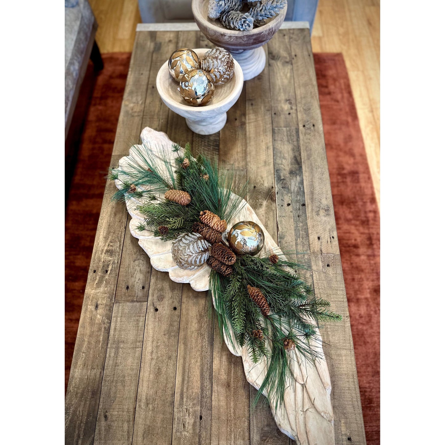 An Angel Wing Wall Art complements the fall-themed wooden coffee table centerpiece with pine branches, brown pinecones, and gold foil ornaments on a wooden leaf tray. The setup includes two matching bowls on one end and rests atop a red-brown area rug over the wooden floor.