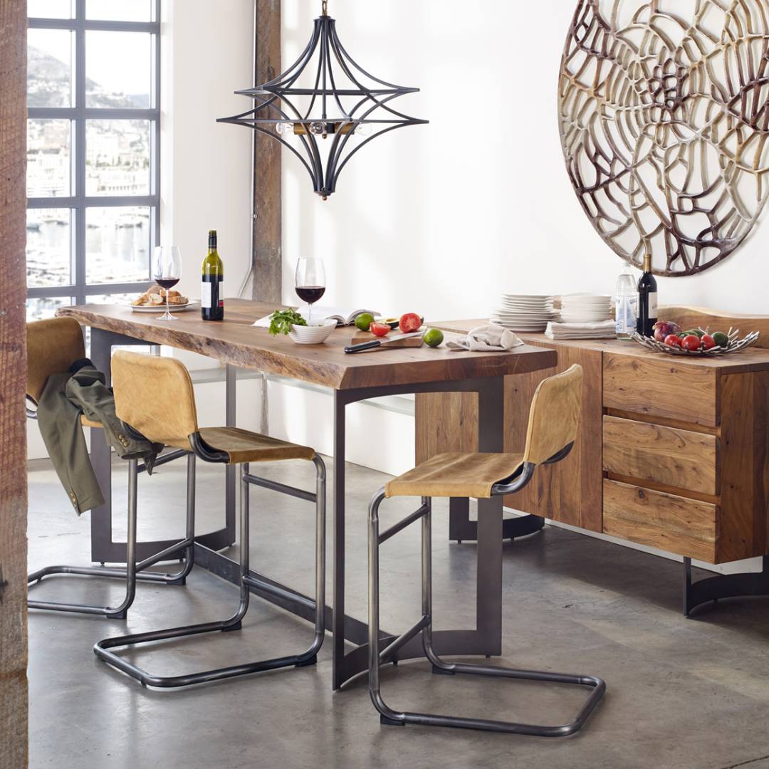 Modern dining room featuring the Bentley Live Edge Counter Table, four wooden chairs with metal legs, and a geometric black chandelier. Wine, glasses, and food are set out as sunlight streams in on wall art and white walls.