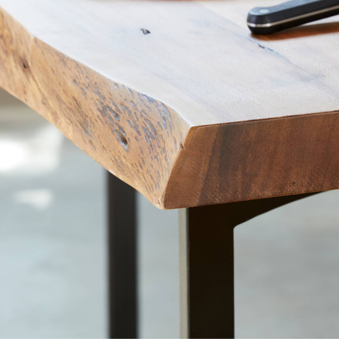 Close-up of the Bentley Live Edge Counter Table's solid acacia wood corner, featuring a live natural edge, visible wood grain, and smooth finish. The tabletop sits on black metal legs; a black object rests on it with a blurred light background.