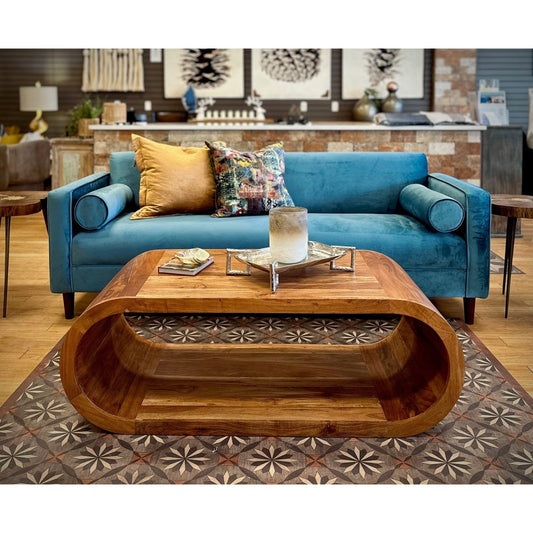 A contemporary living room features a Bench Velvet Blue Sofa adorned with colorful pillows, facing a unique wooden coffee table with floral patterns. The background shows a stylish interior with various decor elements.