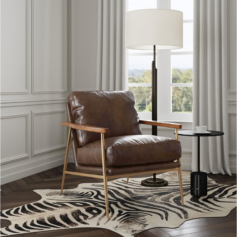 A cozy interior showcases an industrial chic vibe with the Gimbal Leather Accent Chair in brown, featuring wooden armrests. It sits on a zebra-patterned rug over dark wood flooring. Beside it is a small black round table and a tall floor lamp with a white shade. Cream curtains frame windows with greenery views.