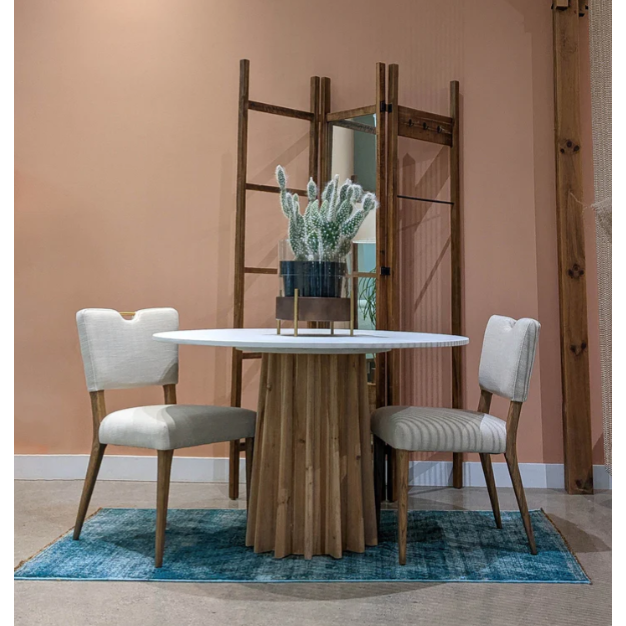 A small round table with a wooden base and white top sits on a blue rug, flanked by two Arden Gold Handle Dining Chairs with light upholstery and wooden legs. A potted cactus decorates the table, set against a peach wall and wooden divider.