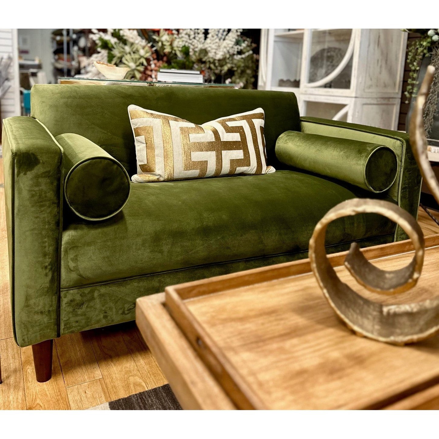 A plush, Bench Velvet Loveseat, Olive Green with two cylindrical side pillows and one decorative geometric-patterned pillow in the center. The loveseat sits in a cozy room setting with a wooden table foreground and floral decor in the background.
