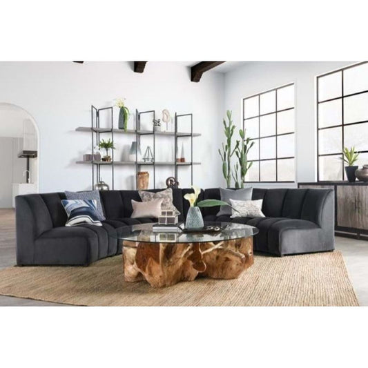 A modern living room with a large, U-shaped Stella Sectional Sofa centered around a unique glass coffee table with a natural wooden base. The room features light walls, large windows, a jute rug, and a metal shelving unit with plants and decor.