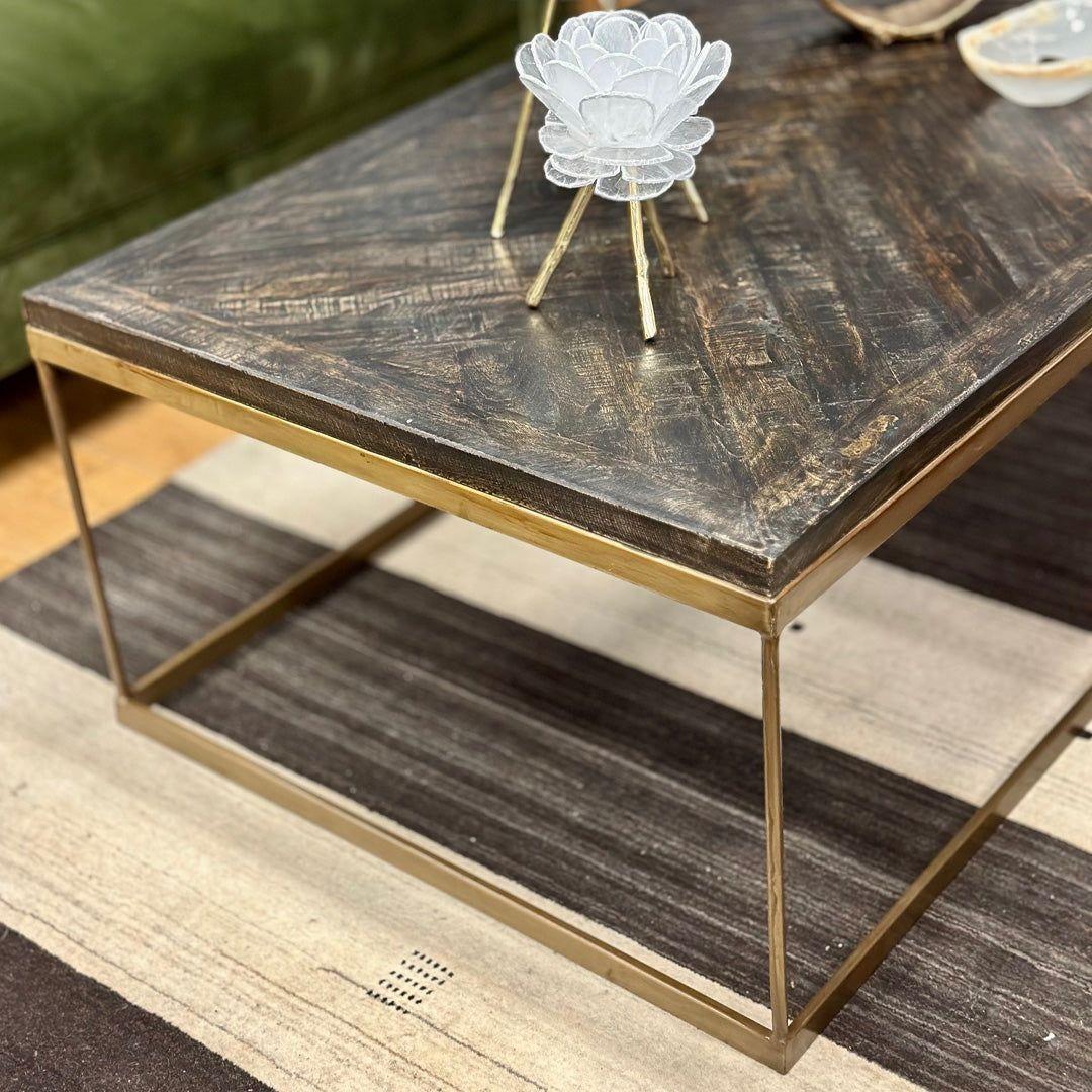 The Parquette Coffee Table, with its rustic charm and dark, textured finish supported by a geometric brass frame, rests on a striped area rug. Atop the table, a white lotus-shaped decorative object draws attention. In the background, part of a green sofa is visible along with a small bowl containing assorted items.