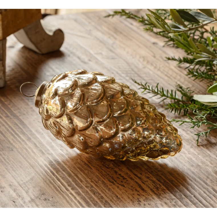 The Burnished Pinecone Ornament, crafted from glass with a golden hue, rests on a rustic wooden surface. Its metallic finish accented by intricate, textured scales exemplifies elegance. To the right, sprigs of green foliage offer a natural touch. The ornament is designed with a small loop at the top for easy hanging.