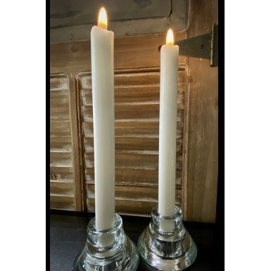 Two white Wax-Dipped Taper Glow Candles are lit, standing side by side in clear glass candle holders. The background features wooden shutters with a rustic finish and a partial metal hinge. The scene has a warm, calming ambiance with a subtle play of light and shadows.