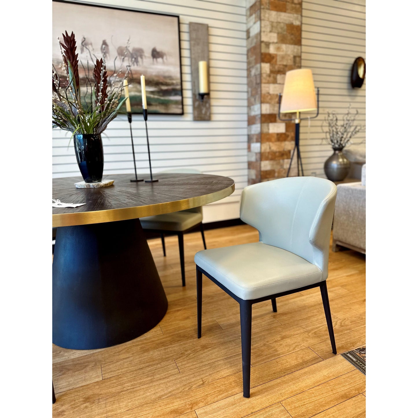 A modern dining room showcases a black round table with a brass edge and the Randolph Dining Chair, which features vegan leather upholstery and black legs. Decor includes a vase with dark flowers, tall candle holders, large artwork, a standing lamp, and textured walls accented with wood and brick.