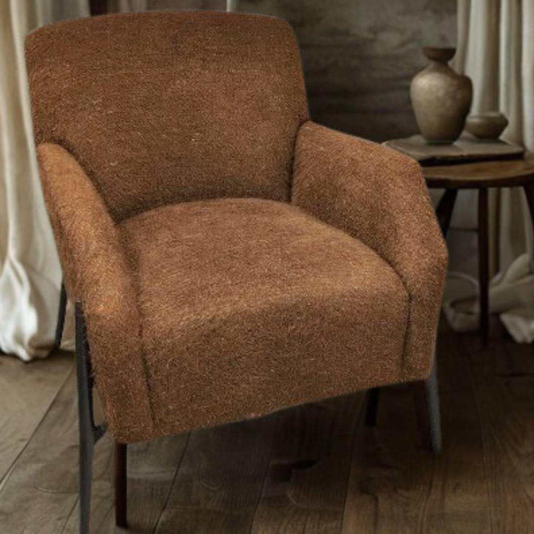 A Sienna Luxe Custom Accent Chair with plush cushions and brown, textured fabric sits on wooden flooring beside a round wood side table holding two ceramic vases, set against beige curtains for a cozy, rustic ambiance.