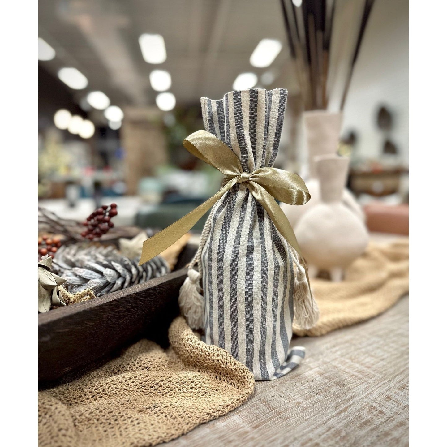 A Wine Bottle Bag with a gray and white striped fabric cover, tied at the neck with a gold ribbon, sits on a wooden surface. Soft lighting and blurred background elements like dried plants and textured fabrics create a cozy, rustic atmosphere.