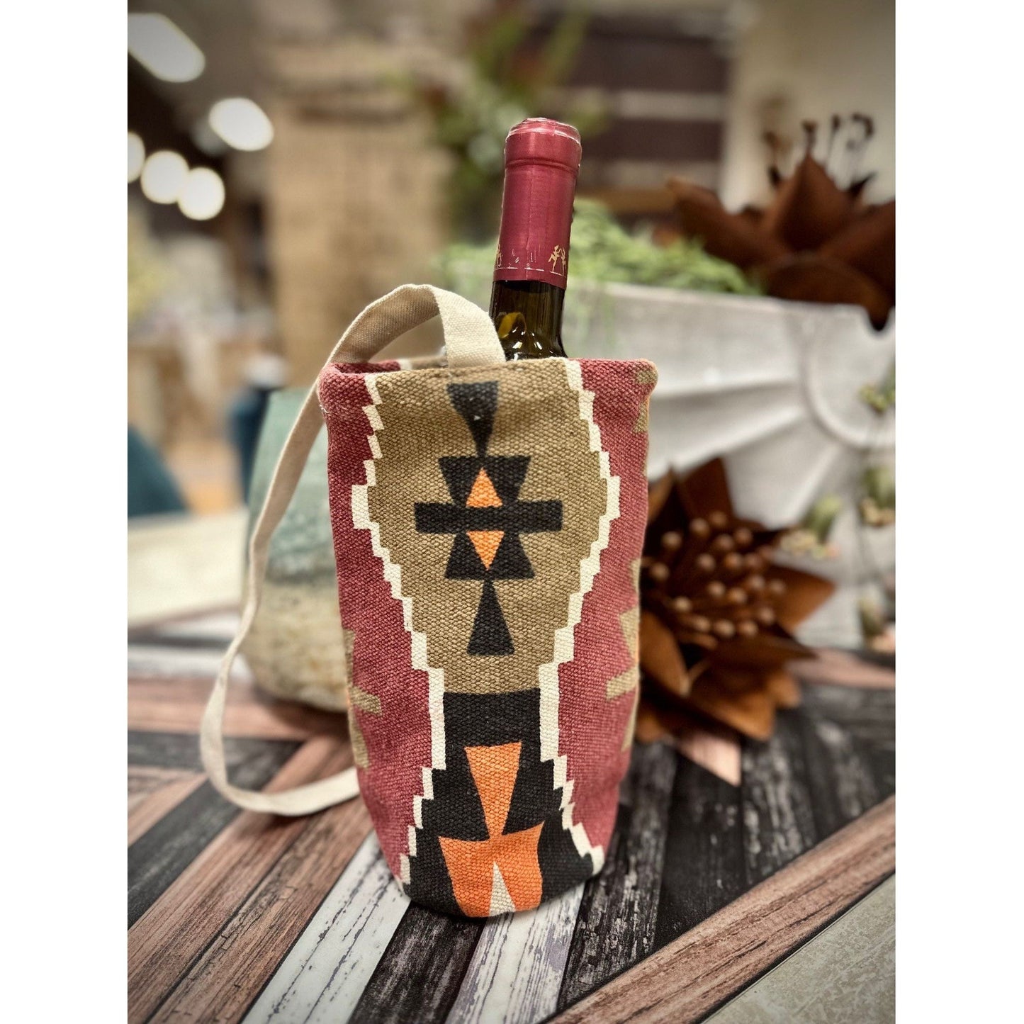 A wine bottle with a red cap is encased in the "Wine Bottle Bag," showcasing a Southwestern geometric design in black, brown, and orange. The bag, featuring a handle, rests on a wooden surface amid blurred soft lighting and nearby plant decor.