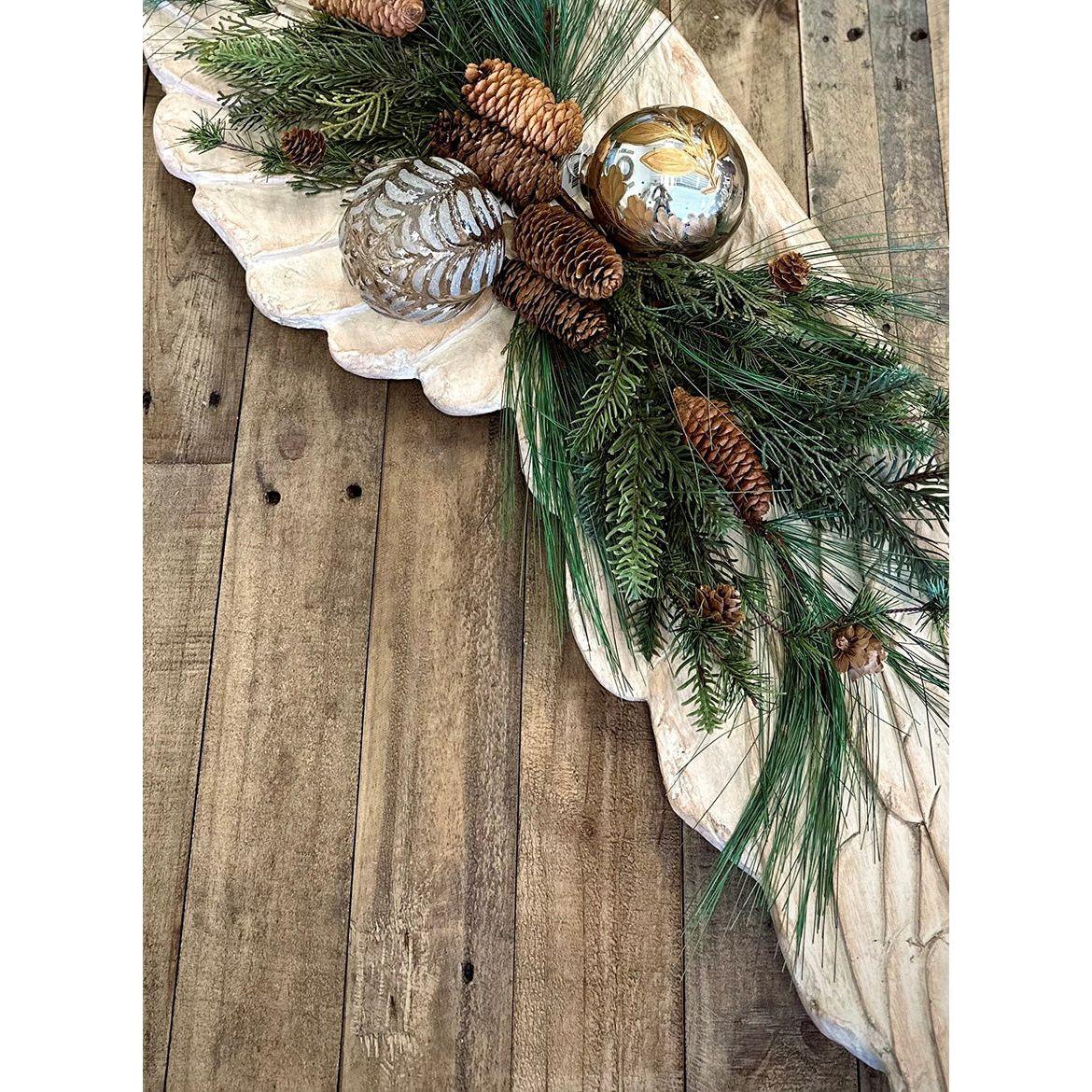 A rustic wooden tray on a table showcases pine branches, pinecones, and decorative gold-silver patterned balls. Accentuated by angel wing wall art, the greenery forms a festive, woodland-themed Christmas centerpiece.