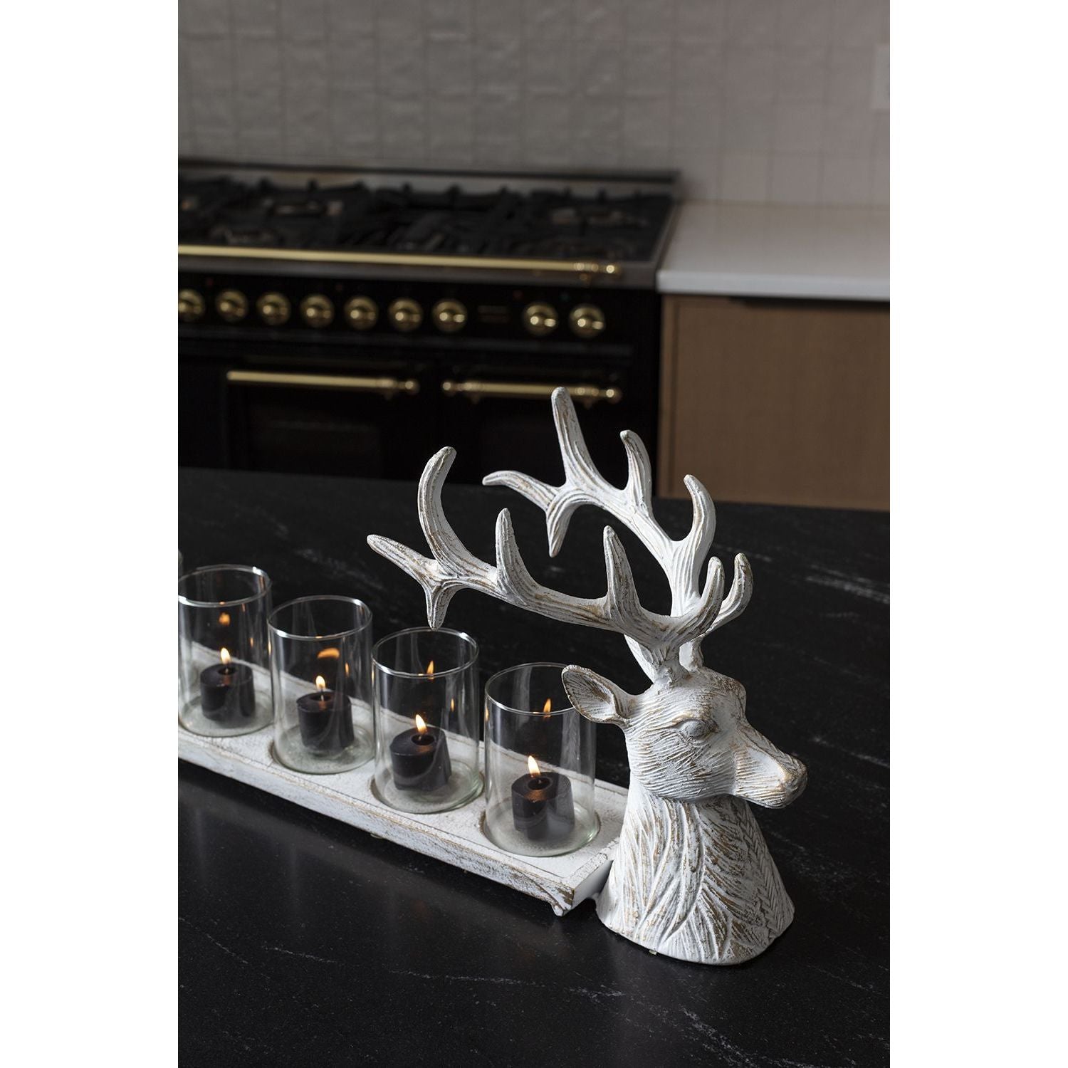 A Reindeer Candleholder is placed on a black countertop. Measuring 35.0 x 6.5 x 11.5, it includes four glass votive holders with lit black candles on a white base. Behind the countertop is a modern kitchen with a black and gold stove and light-colored cabinetry.
