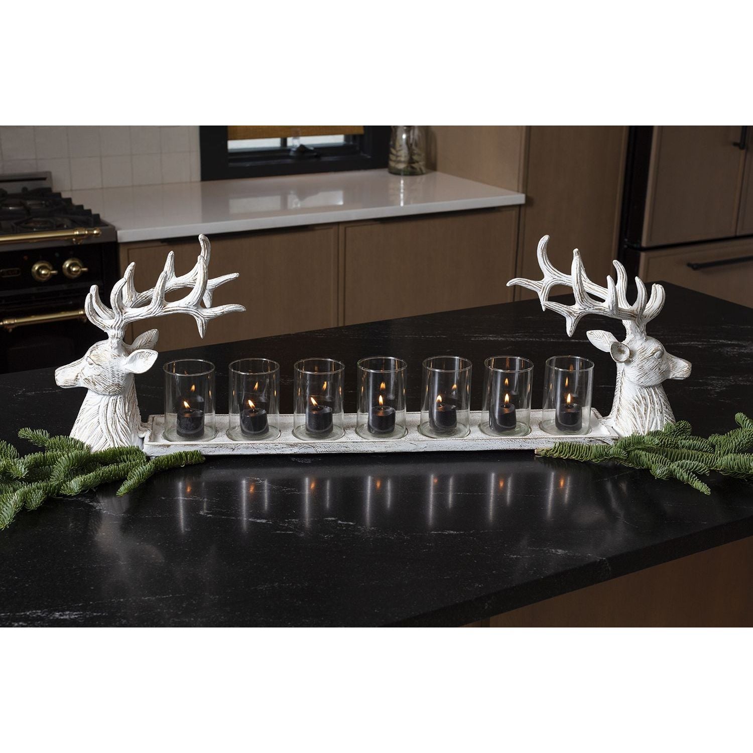 A rectangular black countertop displays a Reindeer Candleholder measuring 35.0 x 6.5 x 11.5 inches. The centerpiece features a row of seven black candleholders with small lit candles inside, mounted on a base with two white reindeer head sculptures on either end, and green artificial pine branches arranged around the base.