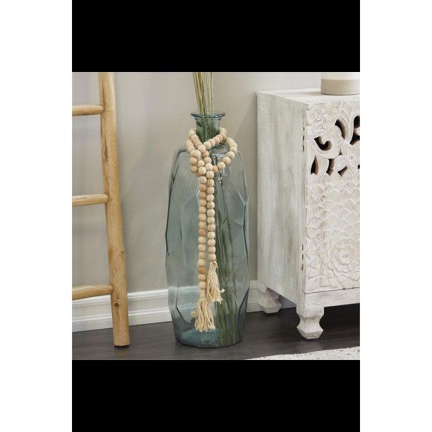 A tall, clear glass vase filled with long, slender stems stands on the floor next to a light-colored wooden cabinet with intricate carvings. Draped over the vase is a Wood Beaded Garland. A wooden ladder leaning against the beige wall is partially visible on the left.