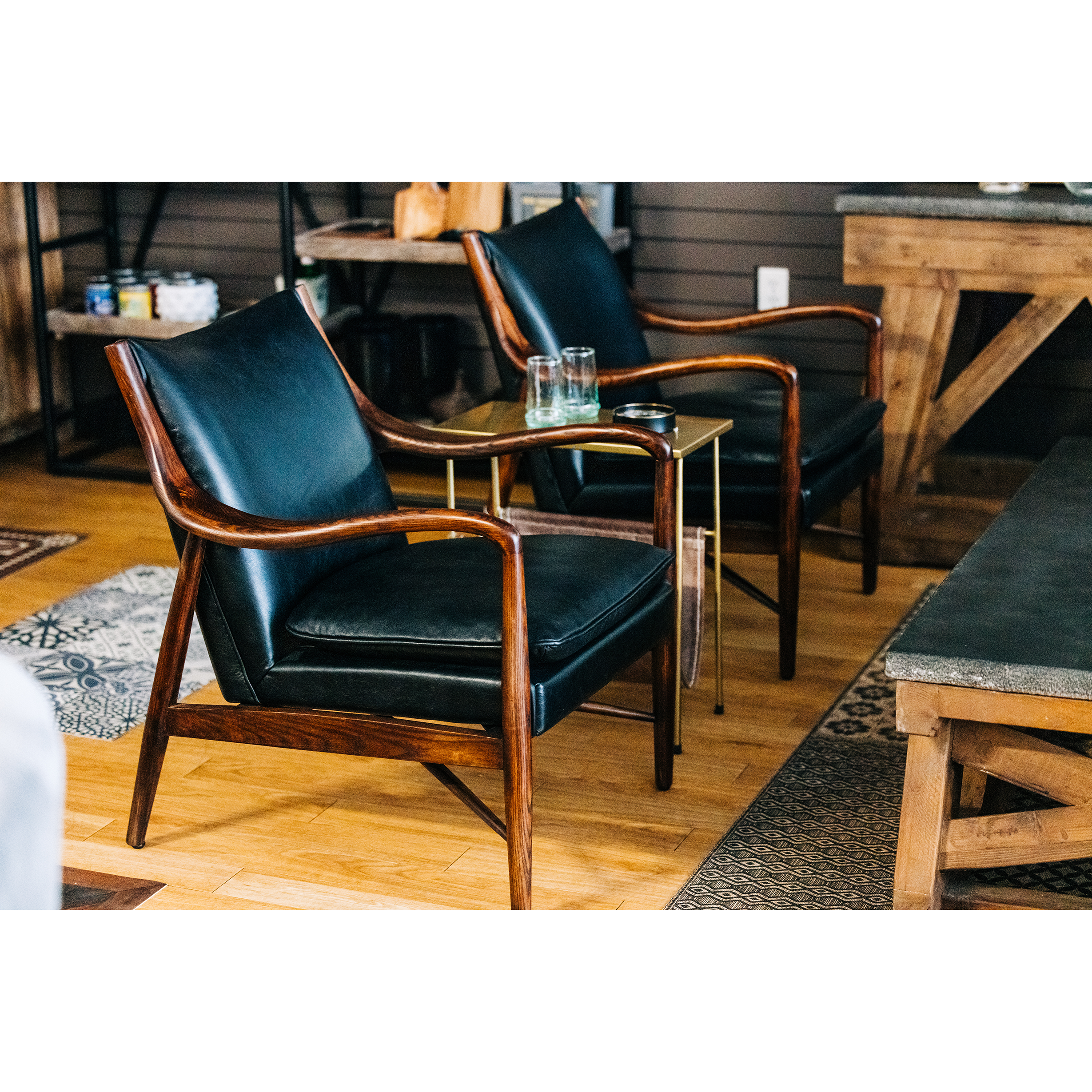 A cozy seating area features two Benson Mid-Century Leather Accent Chairs with walnut frames, positioned around a small round glass table with a beverage on top. The setting has warm wooden flooring and a rustic ambiance.