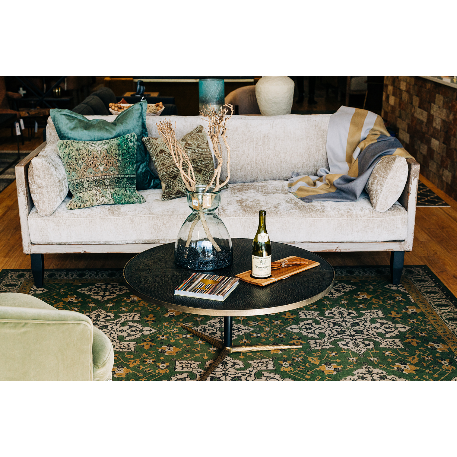 A cozy living room scene with a luxurious Burton Champagne Chenille Sofa adorned with decorative pillows, a round black coffee table featuring a vase with dried twigs, a book, and a wine bottle with a glass on top of a patterned green rug.